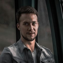 A candid portrait of a man with facial hair wearing a denim jacket, smiling softly.
